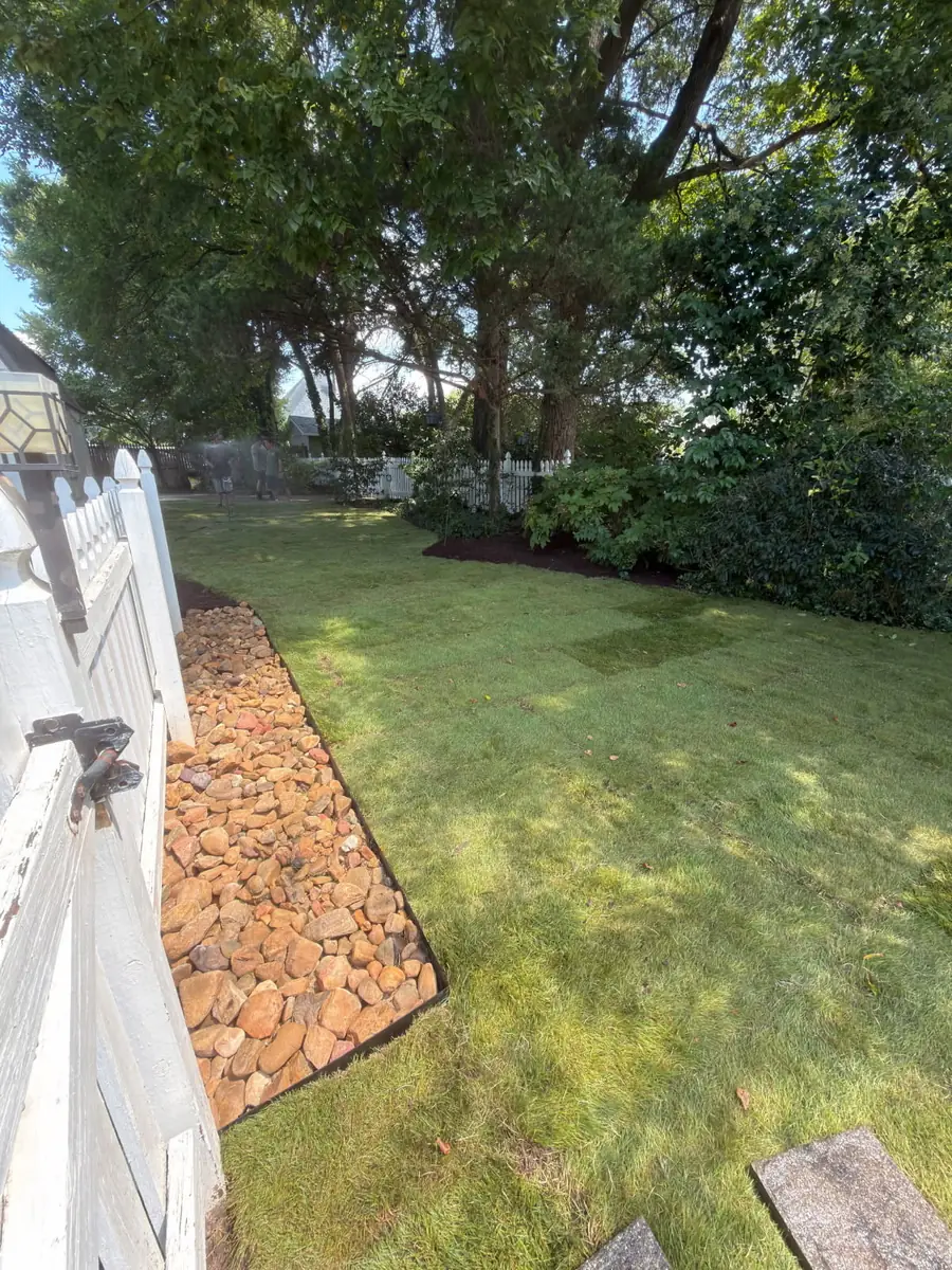 Fresh sod installation in a Concord, NC backyard with river rock border along a white picket fence